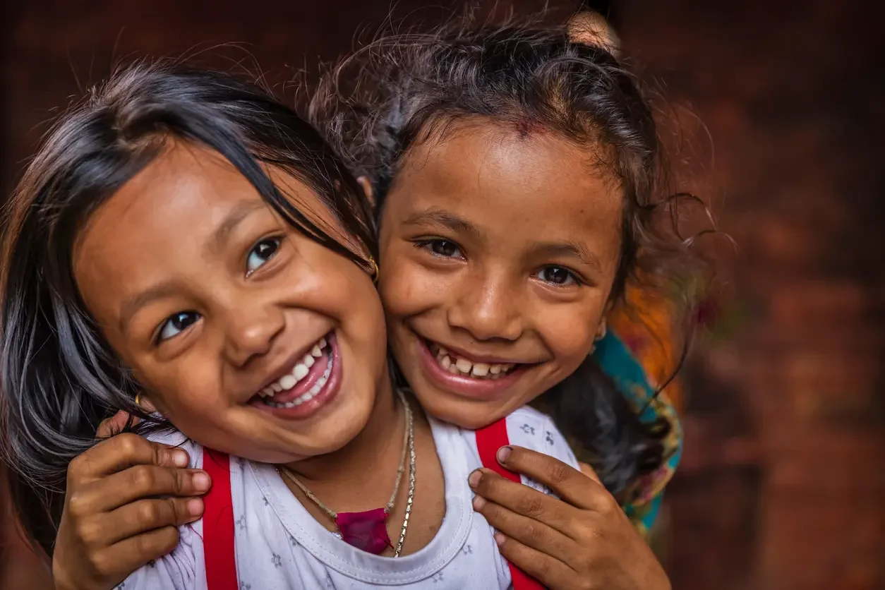 Two young girls smiling and hugging, one with black hair and the other with curly hair, in a close-up outdoor setting.