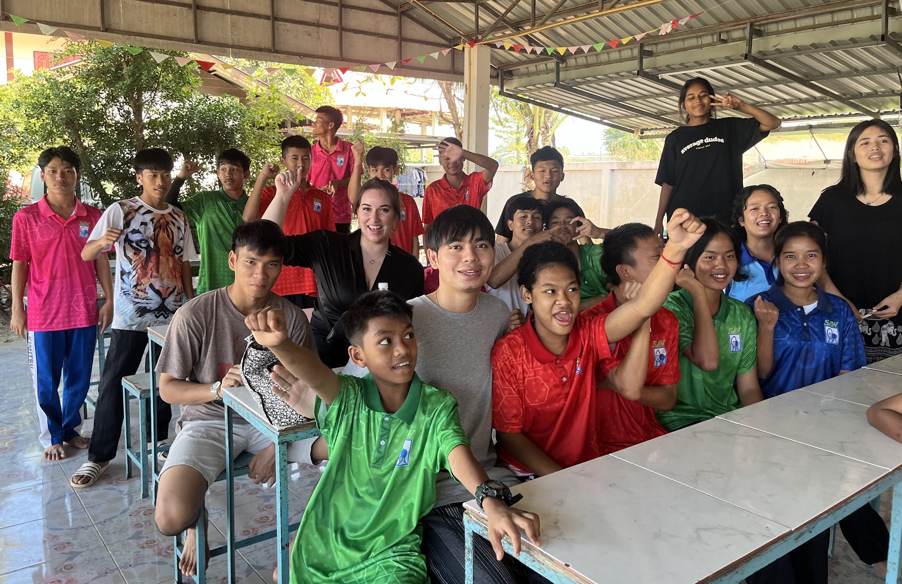 A group of children and teenagers gathered indoors, some standing and some sitting at tables, making gestures like fists and peace signs, smiling and posing for the camera. The setting appears to be a festive or community event with colorful decorations and trees visible outside.