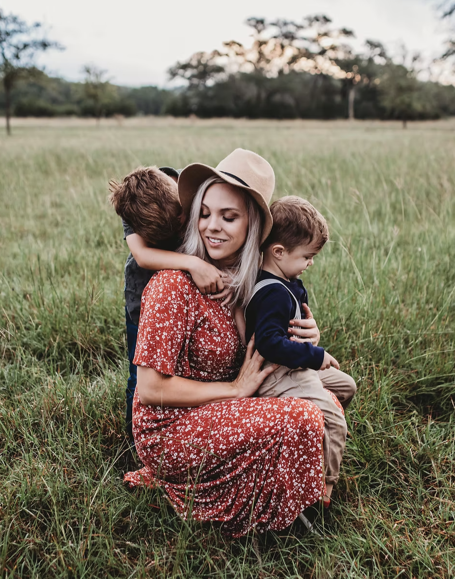 A woman in a red floral dress and beige hat sits in a grassy field, hugging two young boys, one on her lap and the other leaning on her shoulder, during sunset.