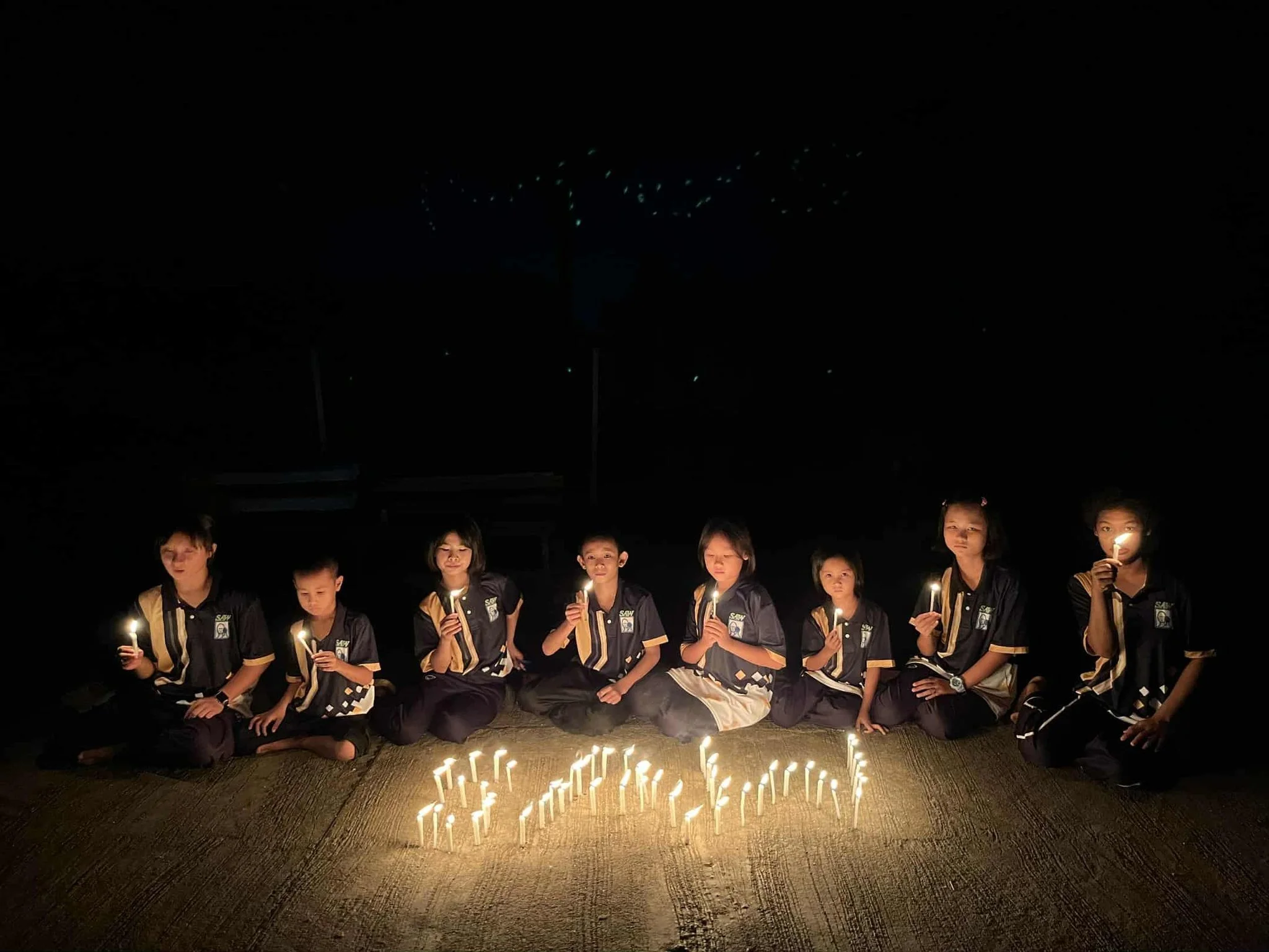 Group of children sitting on the ground at night, holding candles, with lit candles arranged in the shape of a heart on the ground in front of them.