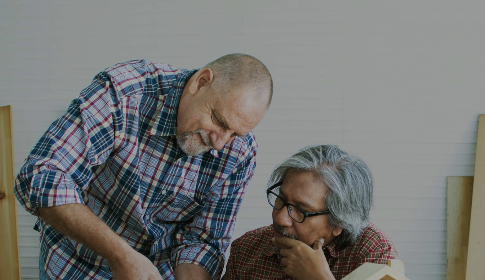Two elderly men in checkered shirts, one with a shaved head and beard, smiling and engaging in a conversation or activity with a third person wearing glasses and gray hair, in a room with light-colored paneled walls.