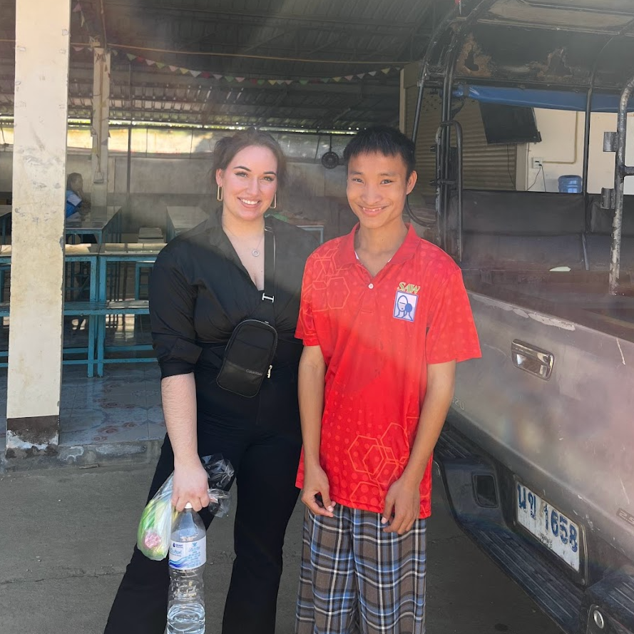 A woman and a boy standing together outdoors, smiling. The woman is holding a water bottle and a small bag of groceries. The boy is wearing a red shirt with a logo and plaid shorts. They are next to a vehicle, with a covered area with tables and chairs in the background.