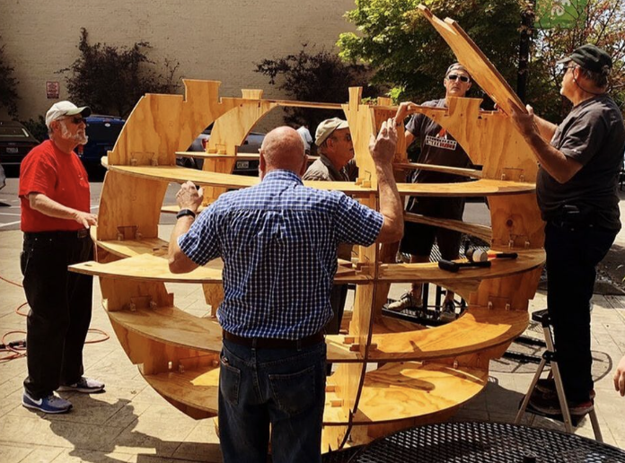 Group of five people assembling or inspecting a large wooden circular shelving unit outdoors on a sunny day.
