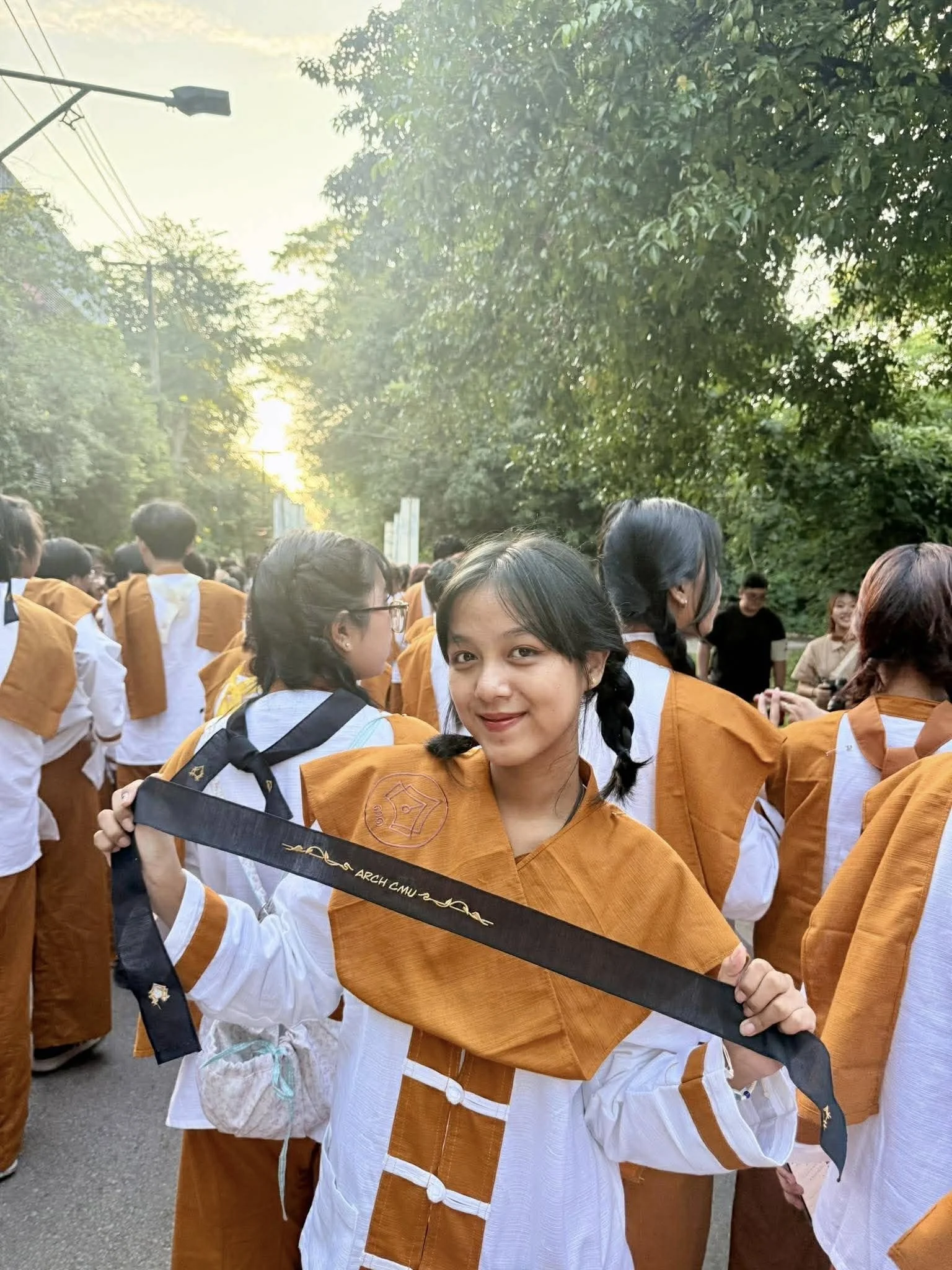 A young woman in a white and orange gown holds a sword with the words 'ARCH CHU' visible, surrounded by a crowd of people dressed similarly near trees at sunset.