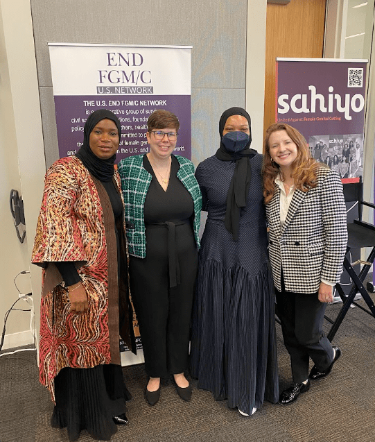 Four women standing together indoors, smiling, with banners in the background related to women's organizations and advocacy.