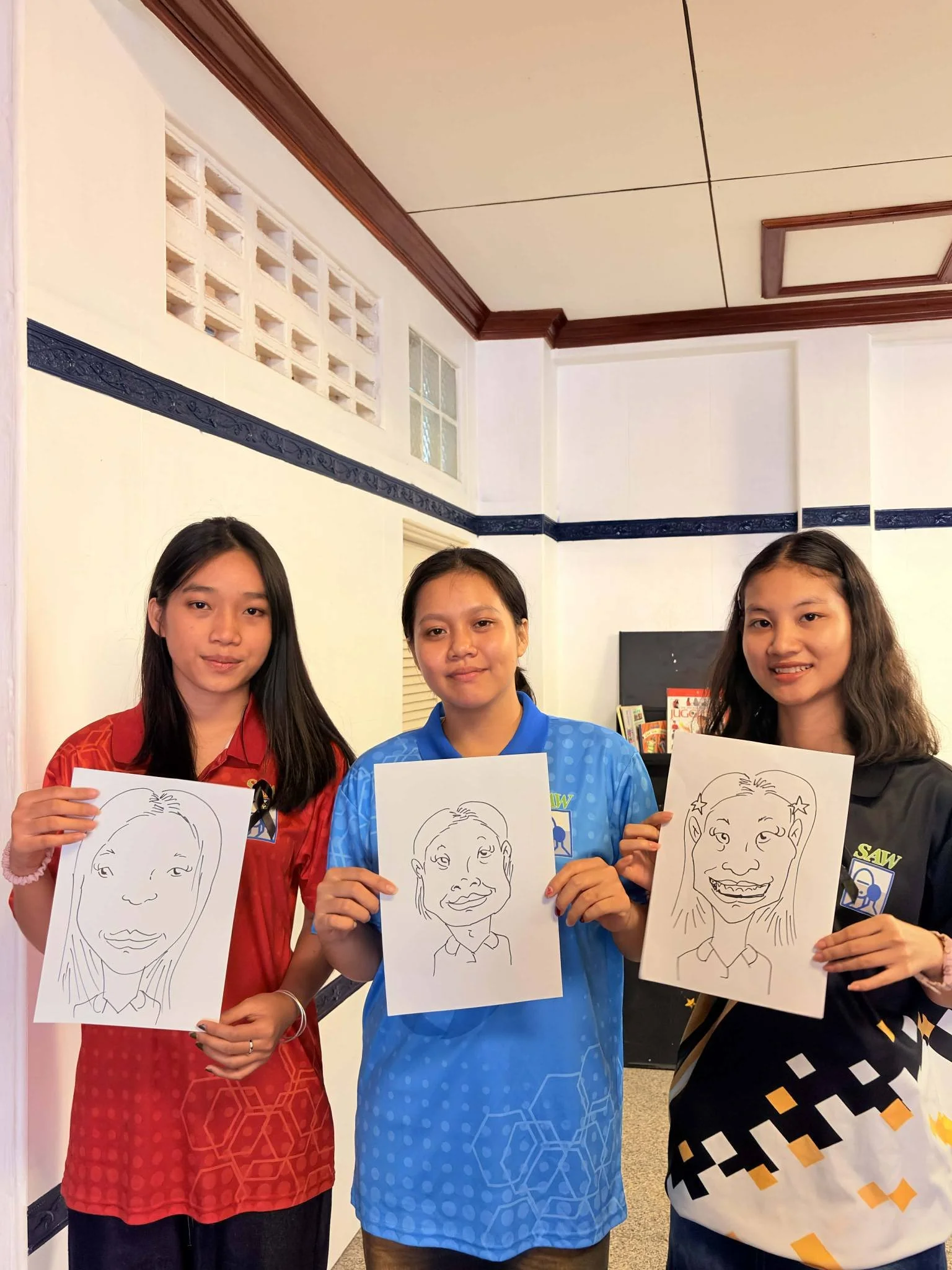 Three young women standing inside a room holding up drawings of exaggerated facial caricatures.