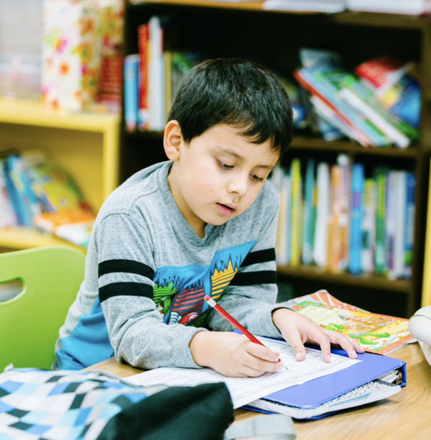 A young boy studying at a desk in a classroom or library, with books and school supplies around him.