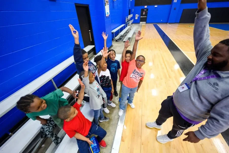 A group of children sitting on gymnasium bleachers raising their hands while a coach or instructor stands in front of them in a gymnasium with a wood floor and blue walls.