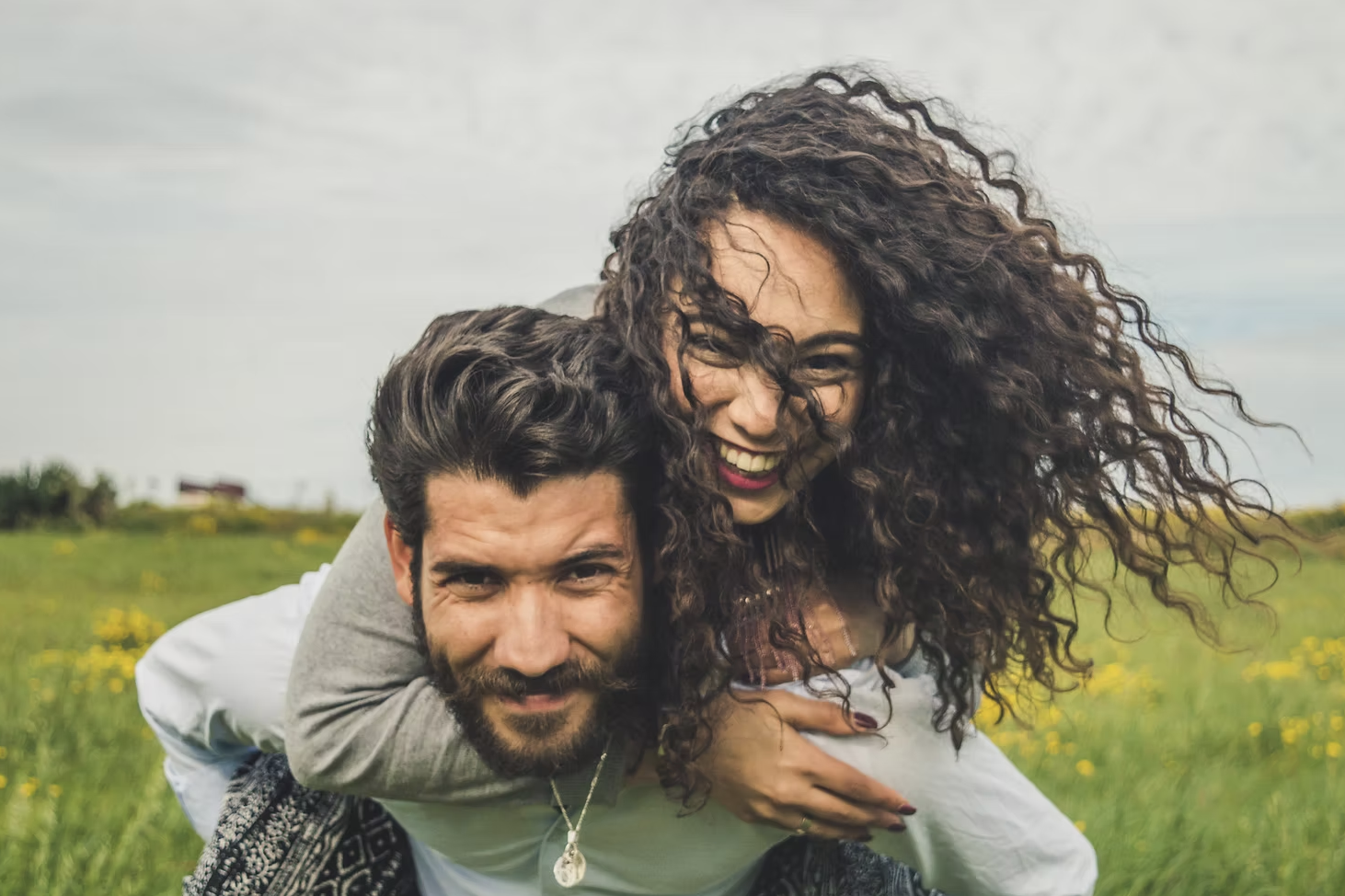 A couple smiling and enjoying in a grassy field with yellow flowers on a cloudy day.