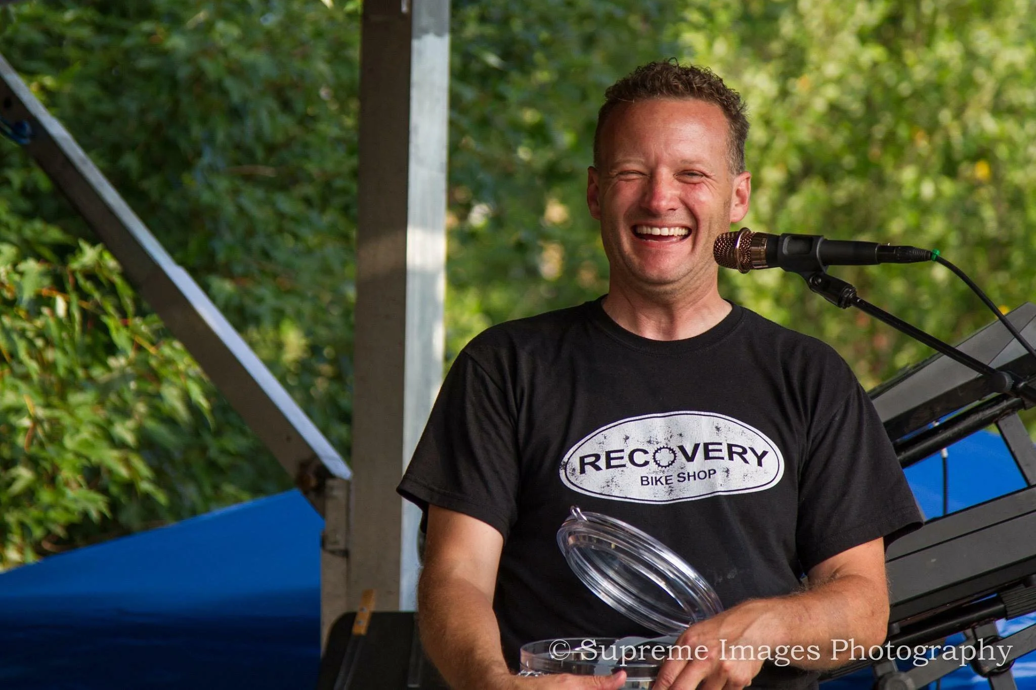 A man with short curly hair smiling and holding a glass, wearing a black T-shirt with the word 'Recovery' and 'Bike Shop' printed on it. He is standing outdoors near a microphone, with green foliage and a structure in the background.