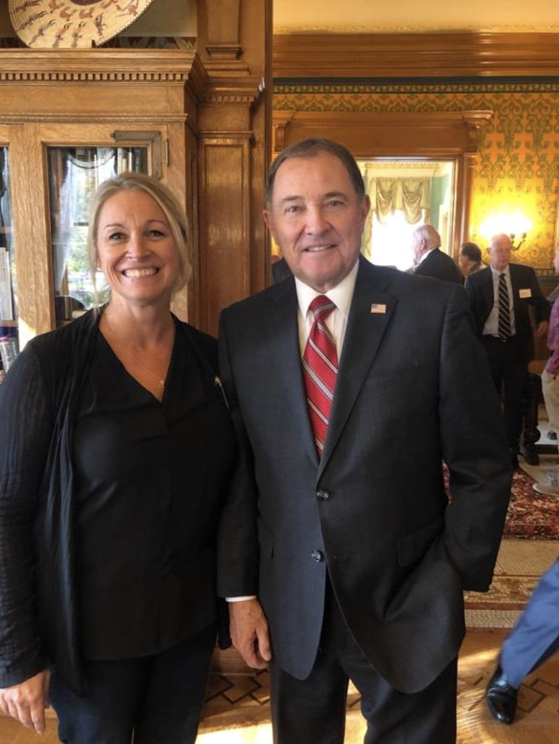 A woman and a man in business attire standing together, both smiling, in an ornate room with wood paneling and yellow patterned wallpaper, during a formal event.