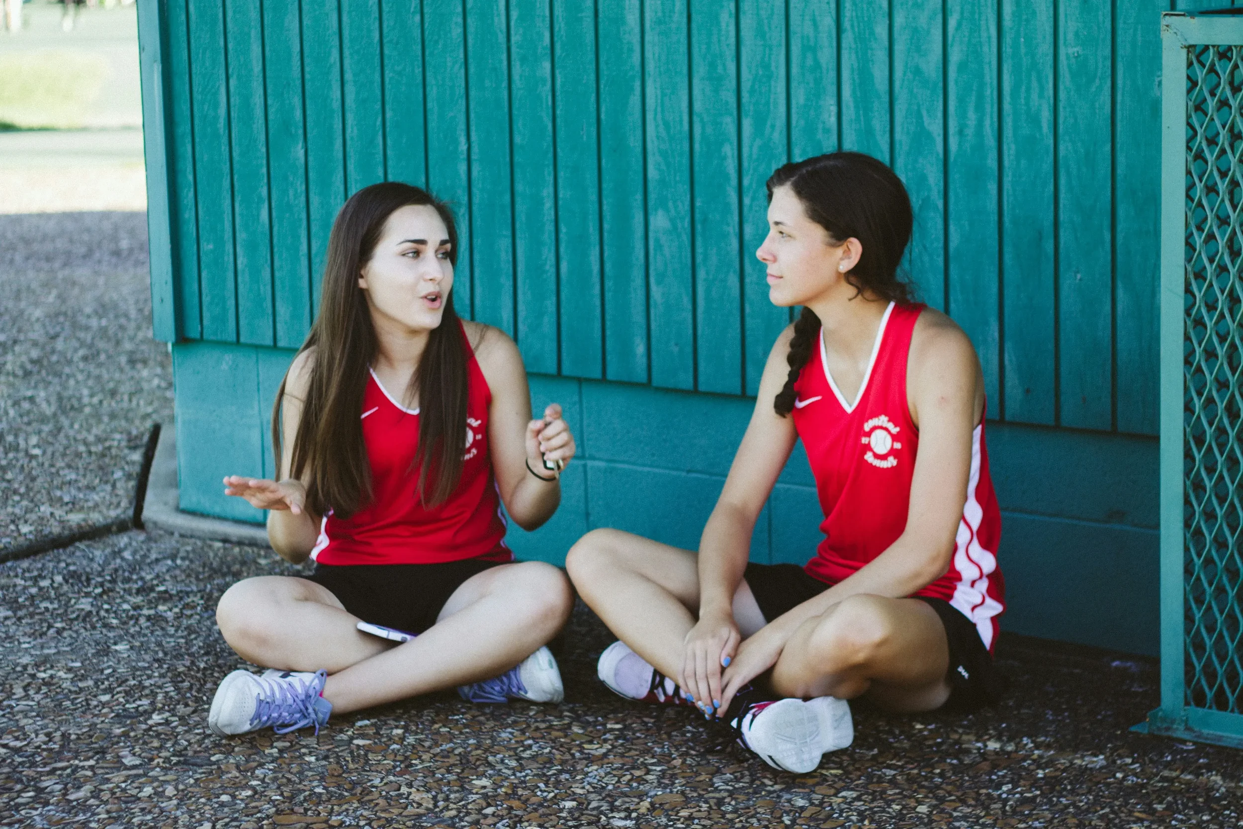 Two young women in red athletic uniforms sitting on the ground outside against a teal wall, having a conversation.