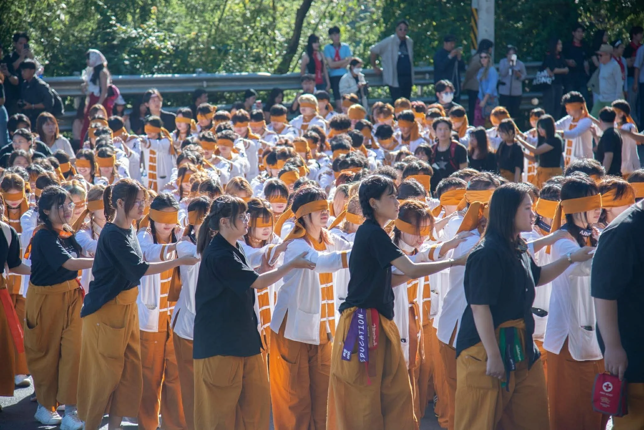 A large group of young people gathered outdoors on a sunny day, many wearing matching outfits with orange and white, and several wearing orange blindfolds, appearing to participate in a coordinated activity or performance.