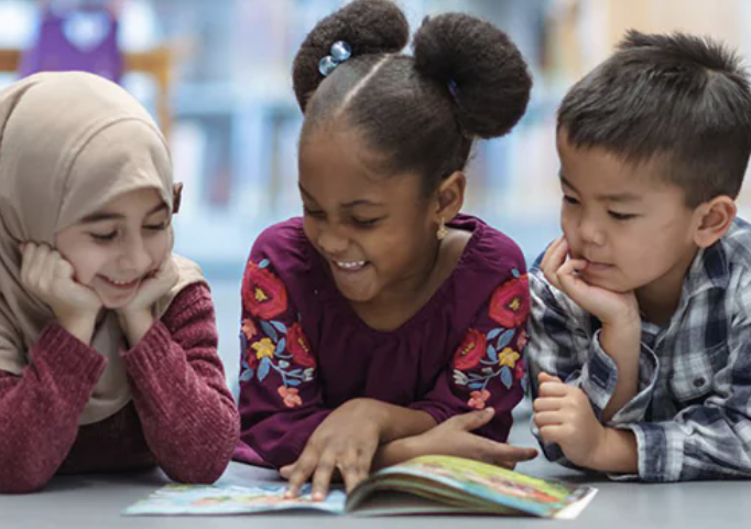 Three children smiling and looking at a book on a table in a classroom.
