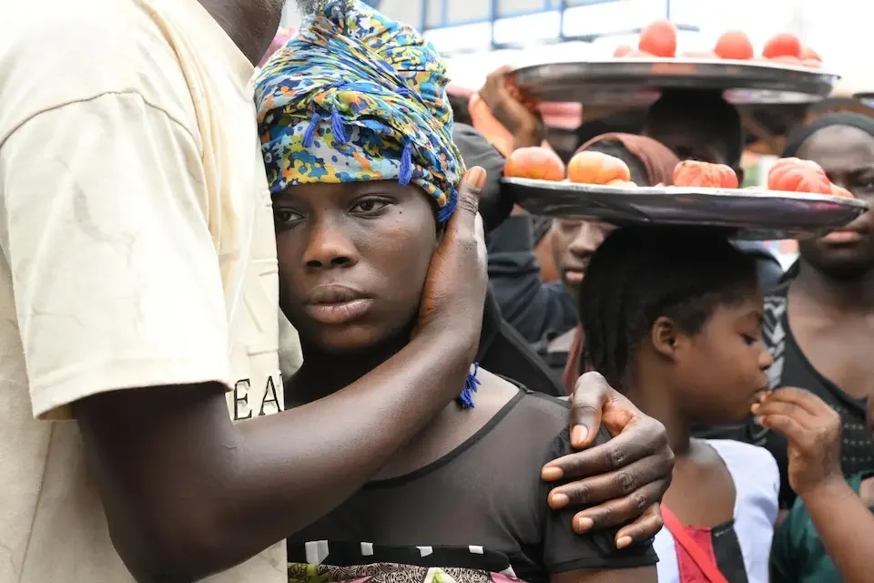A young woman with a headscarf being comforted, with other women in the background holding trays of fruit.