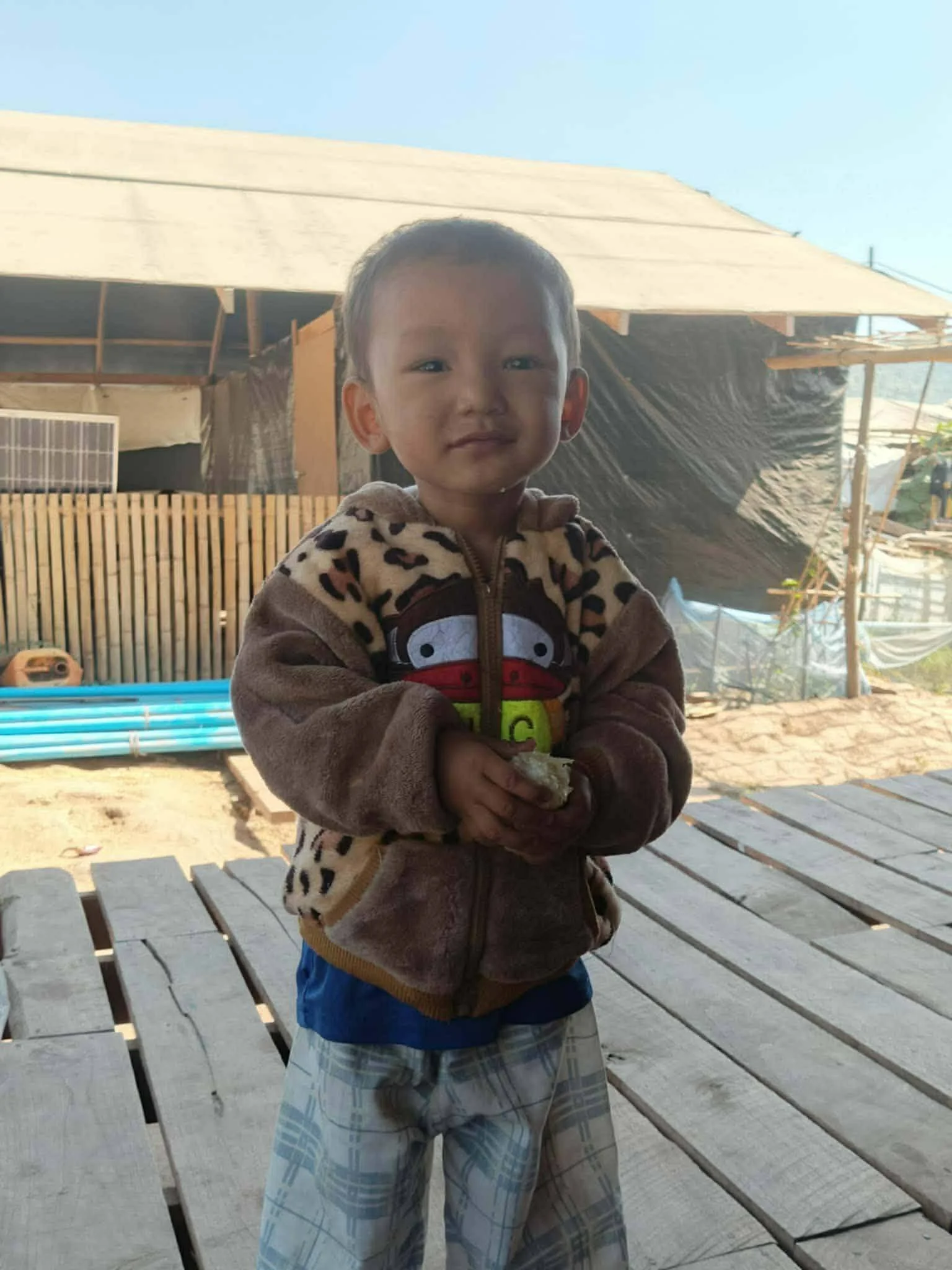 Young boy standing outdoors, wearing a plush hoodie with a cartoon character on it. He has short hair and is holding something in his hands. Background shows a wooden platform, a fence, and a construction site with scaffolding and building materials under a clear sky.