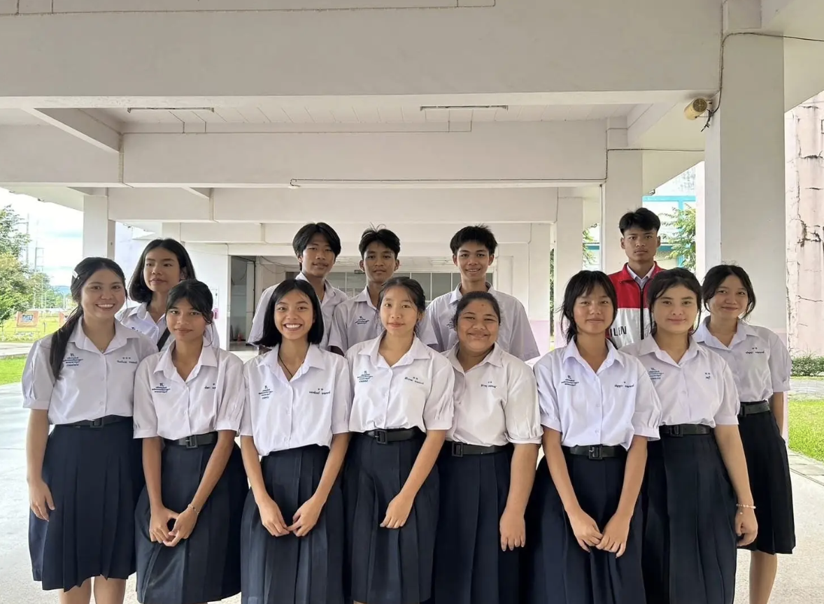 Group of teenage students in school uniforms standing outdoors under a covered area, smiling at the camera.