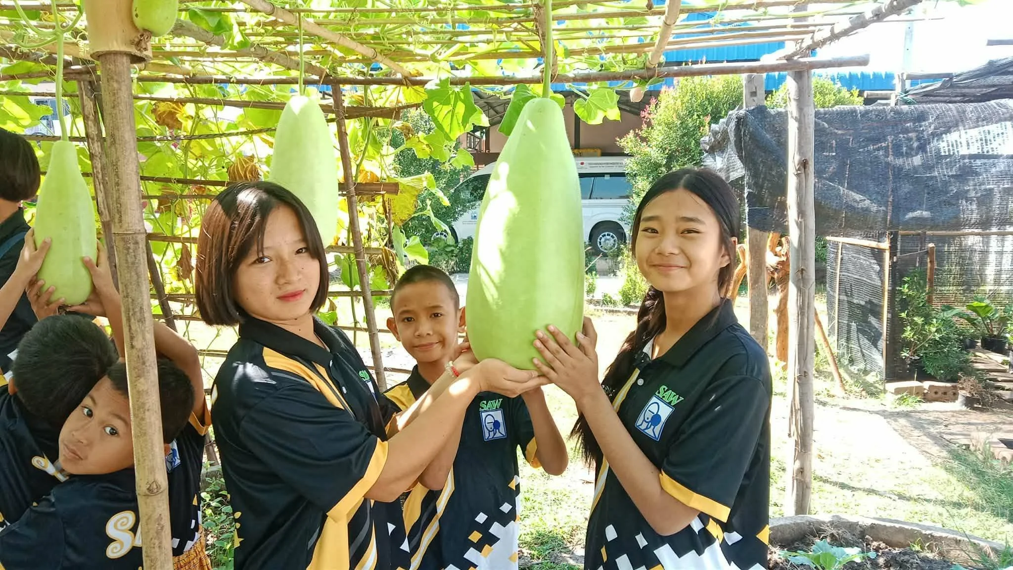 Group of children and two women holding large green gourds in a garden or farm setting.