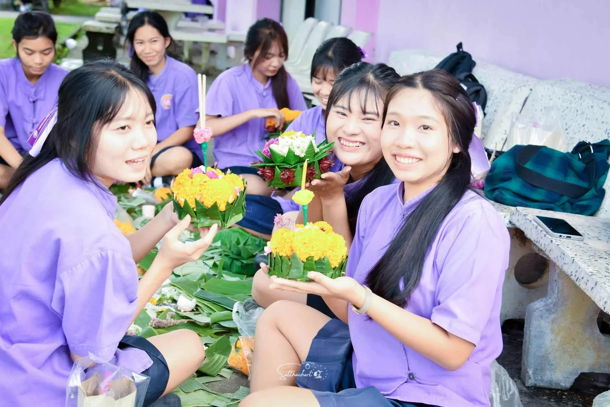A group of young women in purple shirts sitting outdoors around a table, holding colorful floral arrangements made of marigolds, roses, and green leaves for a celebration or festival.