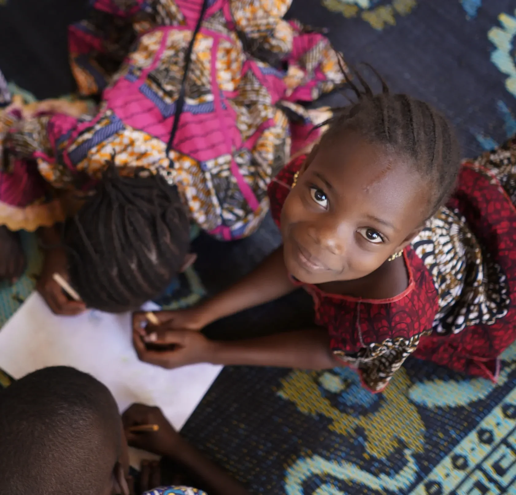 A young girl looking up at the camera while people around her sit on a colorful patterned carpet.