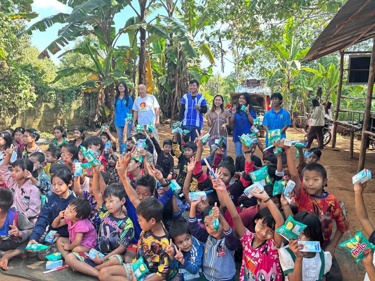 Group of children sitting on the ground holding up snack packages, with adults standing behind them in a tropical outdoor setting with banana trees and a small building.