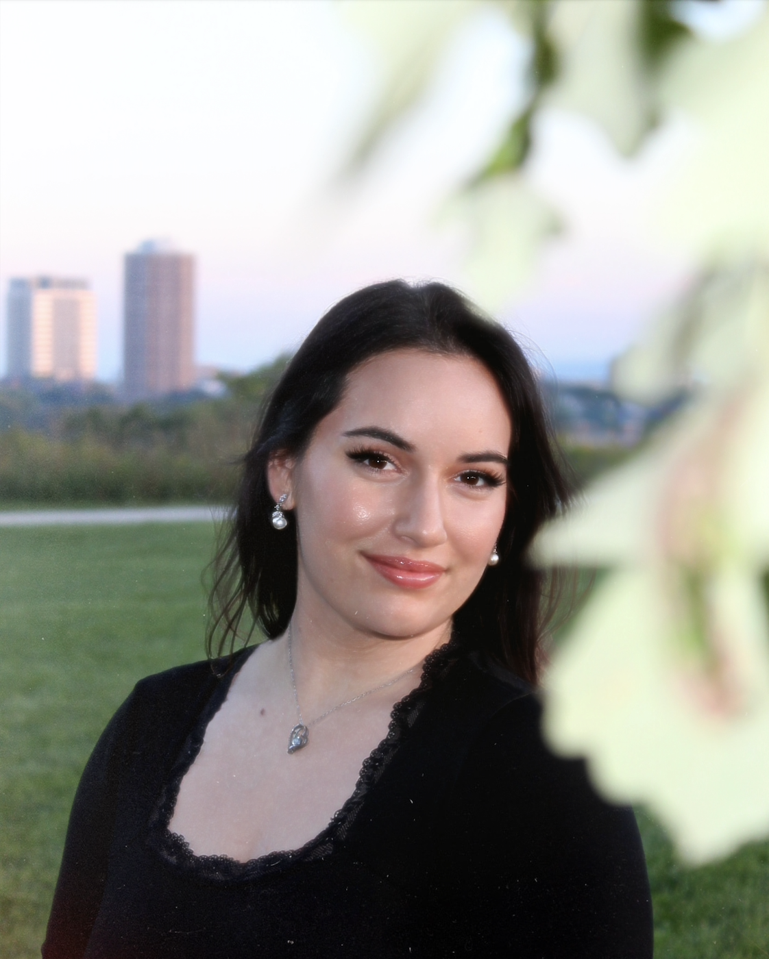 A woman with dark hair, wearing pearl earrings and a necklace, smiling outdoors with a city skyline and green park in the background.
