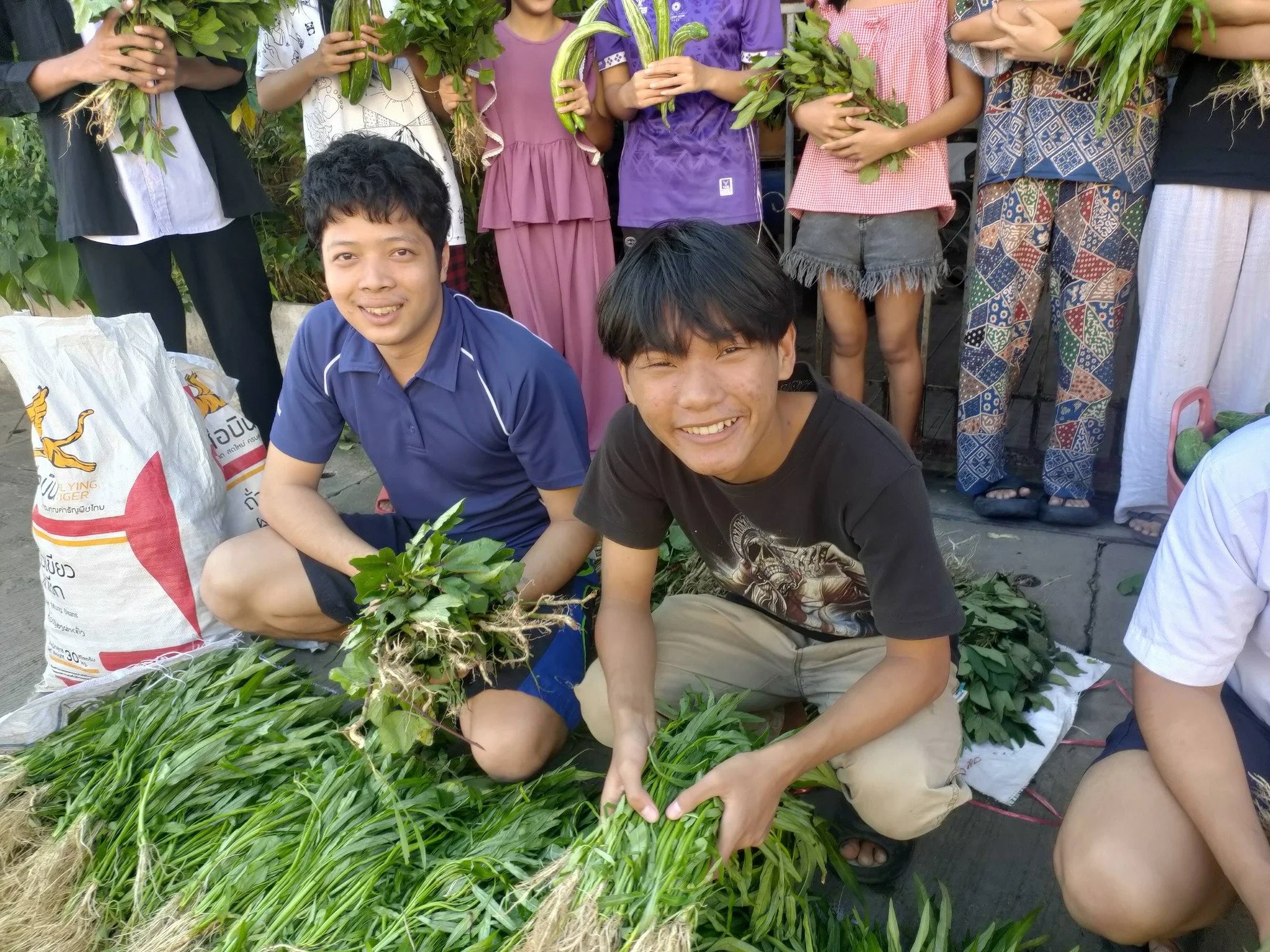 Group of people, mostly children, gathered outdoors holding bunches of green leafy plants and herbs, with some sitting on the ground and others standing.
