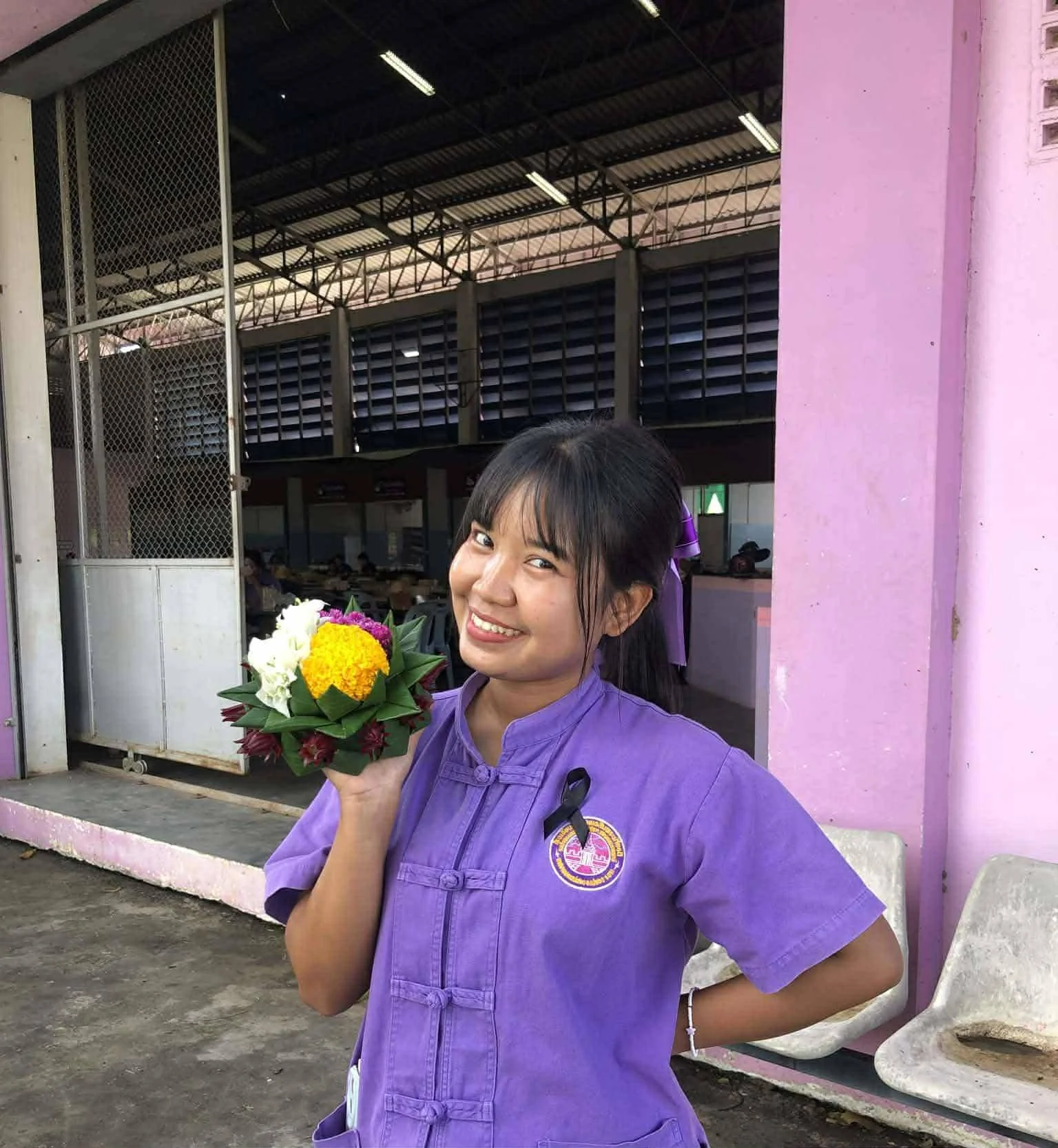 Young girl in purple uniform smiling and holding a colorful flower bouquet, standing outdoors near a pink wall with empty chairs and a building in the background.