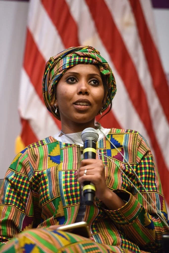 A woman wearing traditional African Kente cloth attire and head wrap, speaking into a microphone at an event with the American flag in the background.