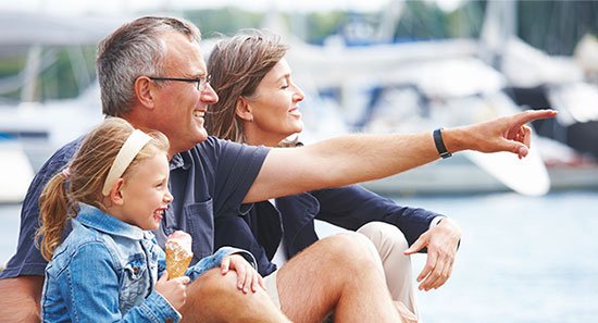 A family of three sitting by the water at a marina, with boats in the background. The father is pointing at something in the distance, and they are smiling and enjoying their time together.
