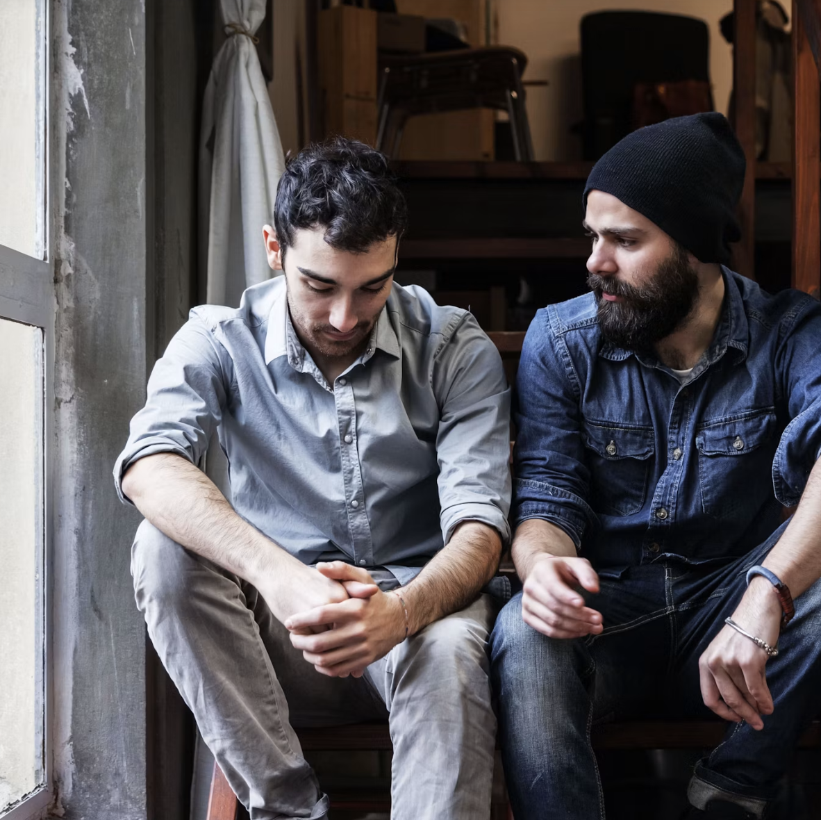 Two men sitting on a bench near a window, one with a gray shirt and the other with a dark beanie and denim jacket, engaging in a serious conversation.