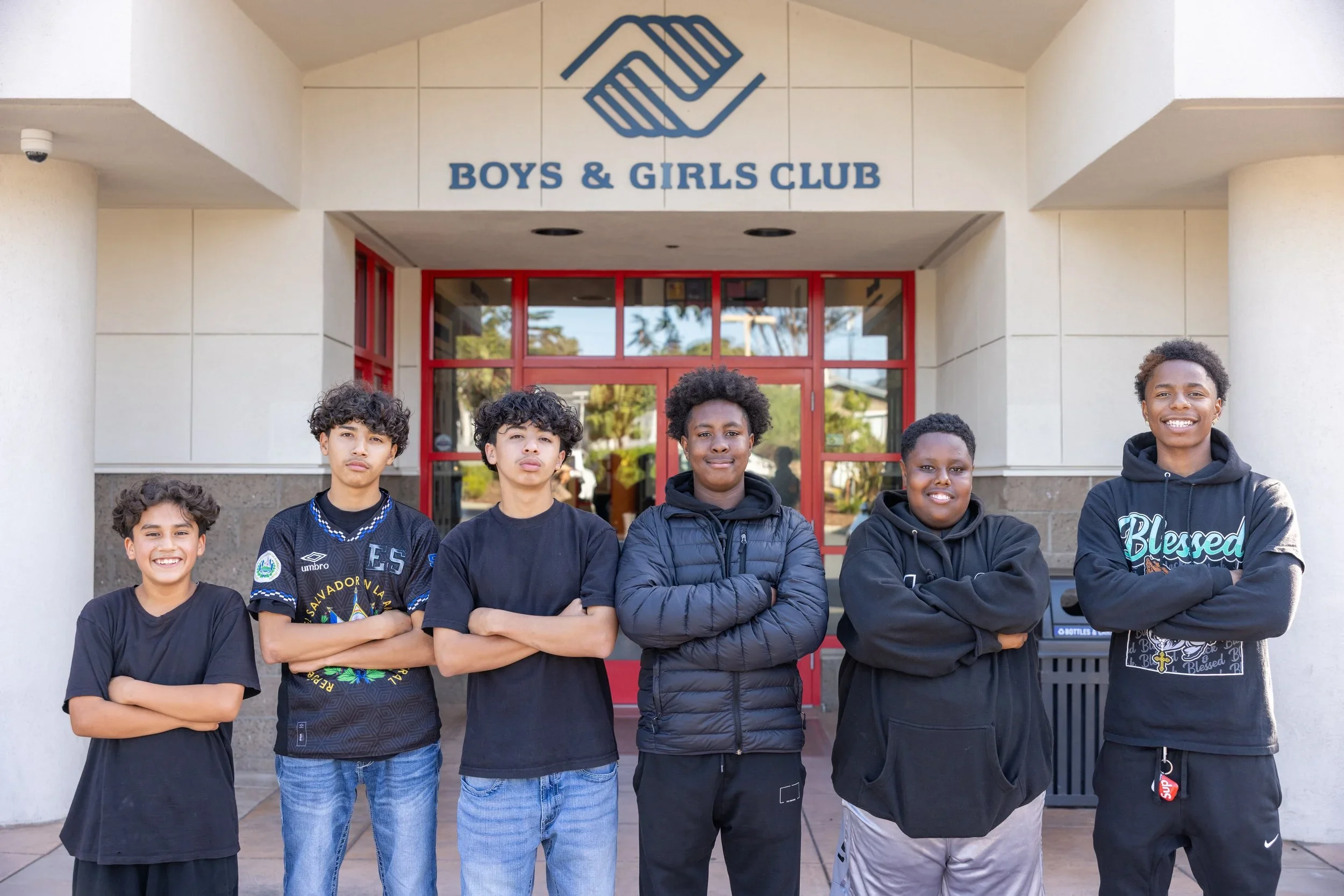 Group of six boys standing outside Boys & Girls Club in front of a red door. They have crossed their arms and are smiling or serious.