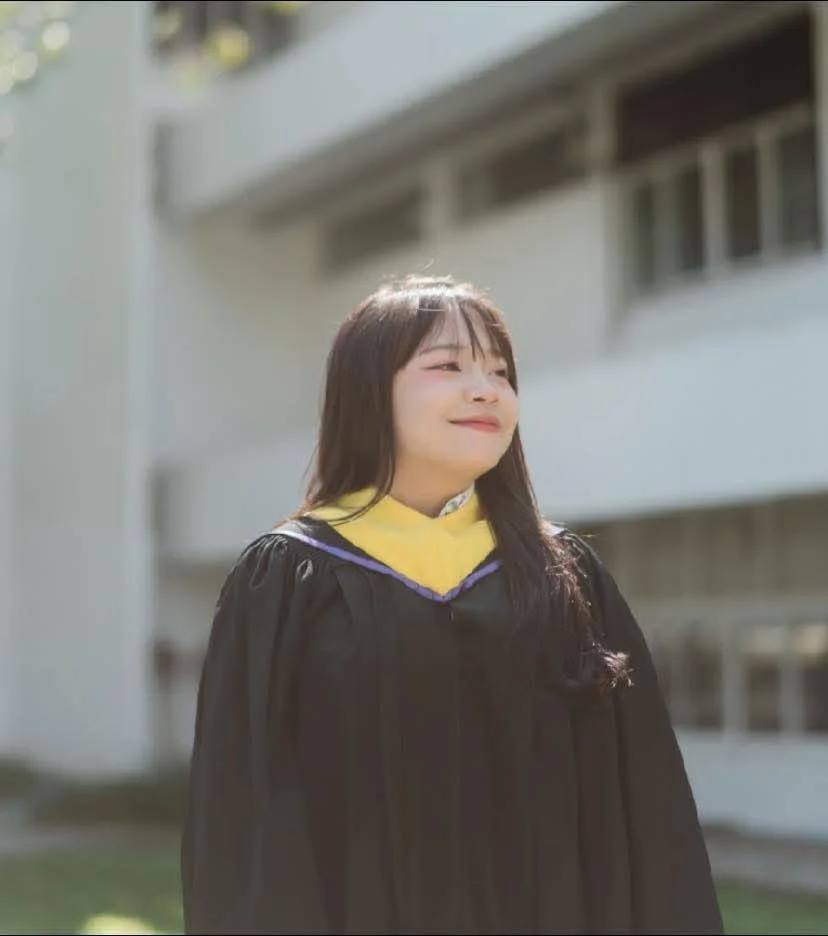 A woman in a graduation gown and cap standing outdoors in front of a building, smiling slightly with her eyes closed.