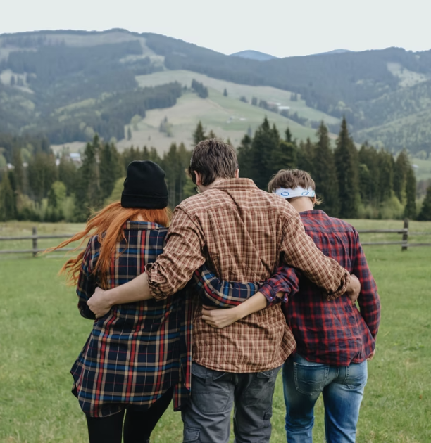 Three friends with arms around each other's shoulders walking across a grassy field with mountains in the background.