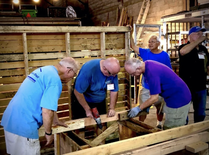 Four men working together on a woodworking project inside a workshop, assembling a wooden structure.