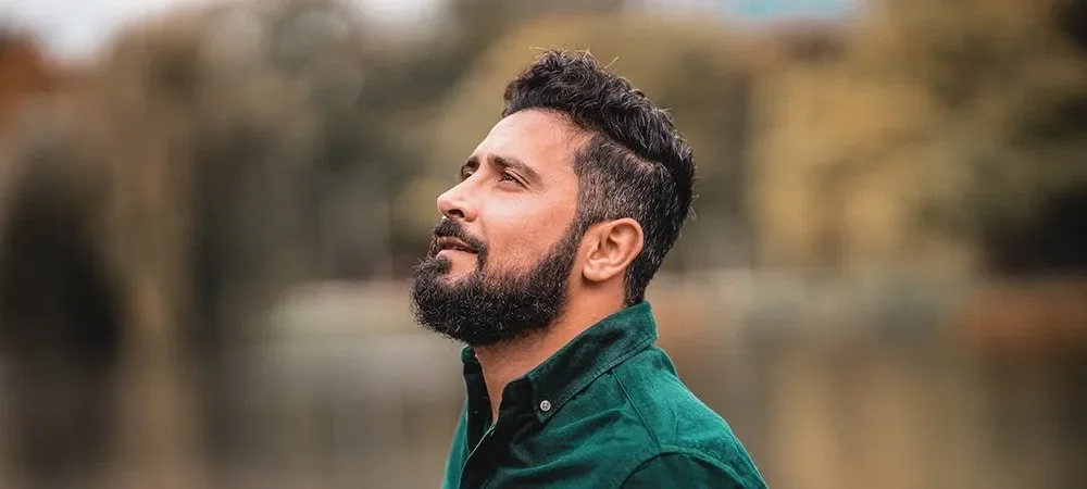 A man with a beard and short dark curly hair, wearing a green shirt, looking upwards outdoors with blurred trees in the background.