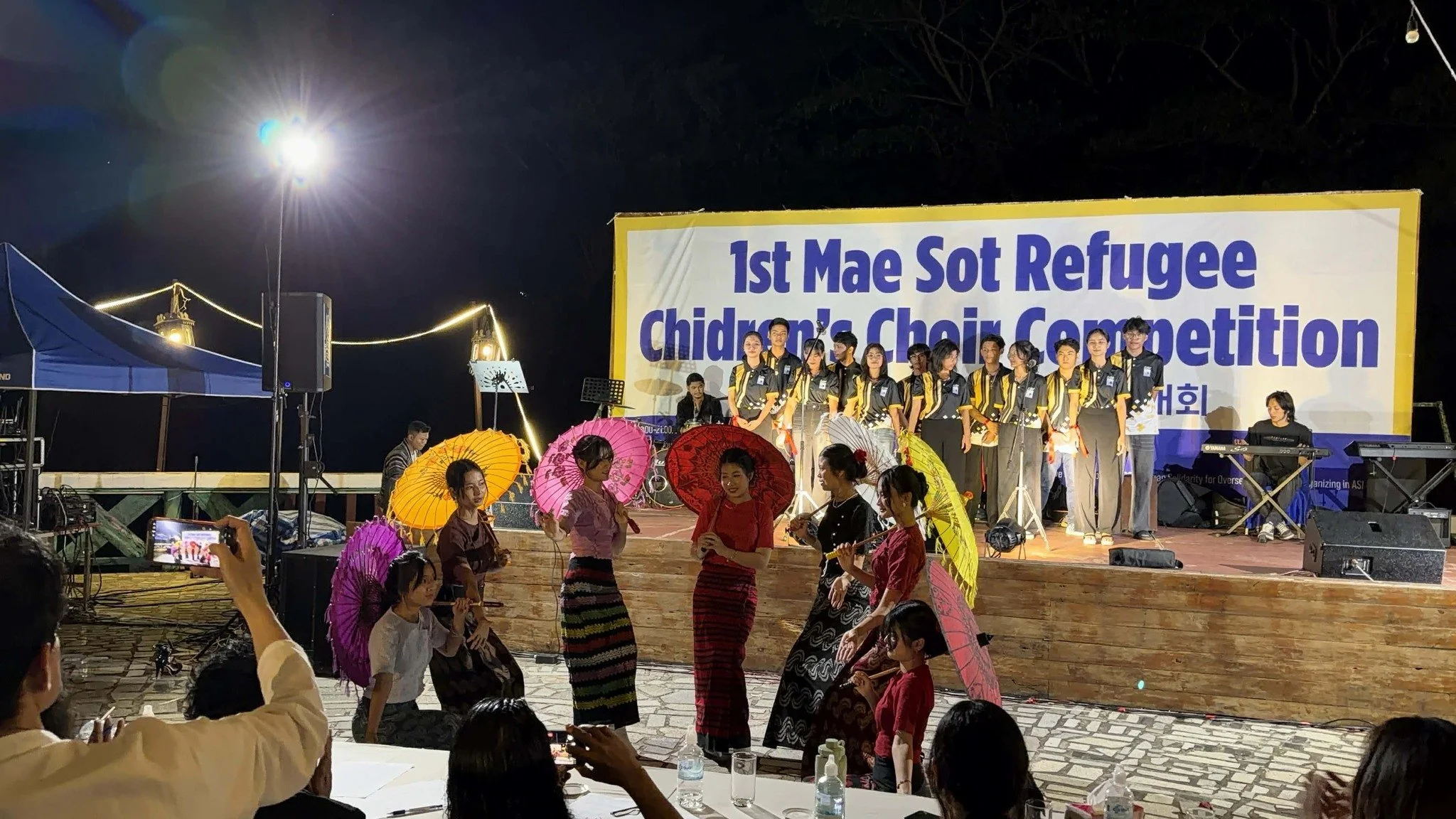 Nighttime cultural event with people holding umbrellas, performers on stage, and a large banner reading '1st Mae Sot Refugee Children’s Choir Competition'