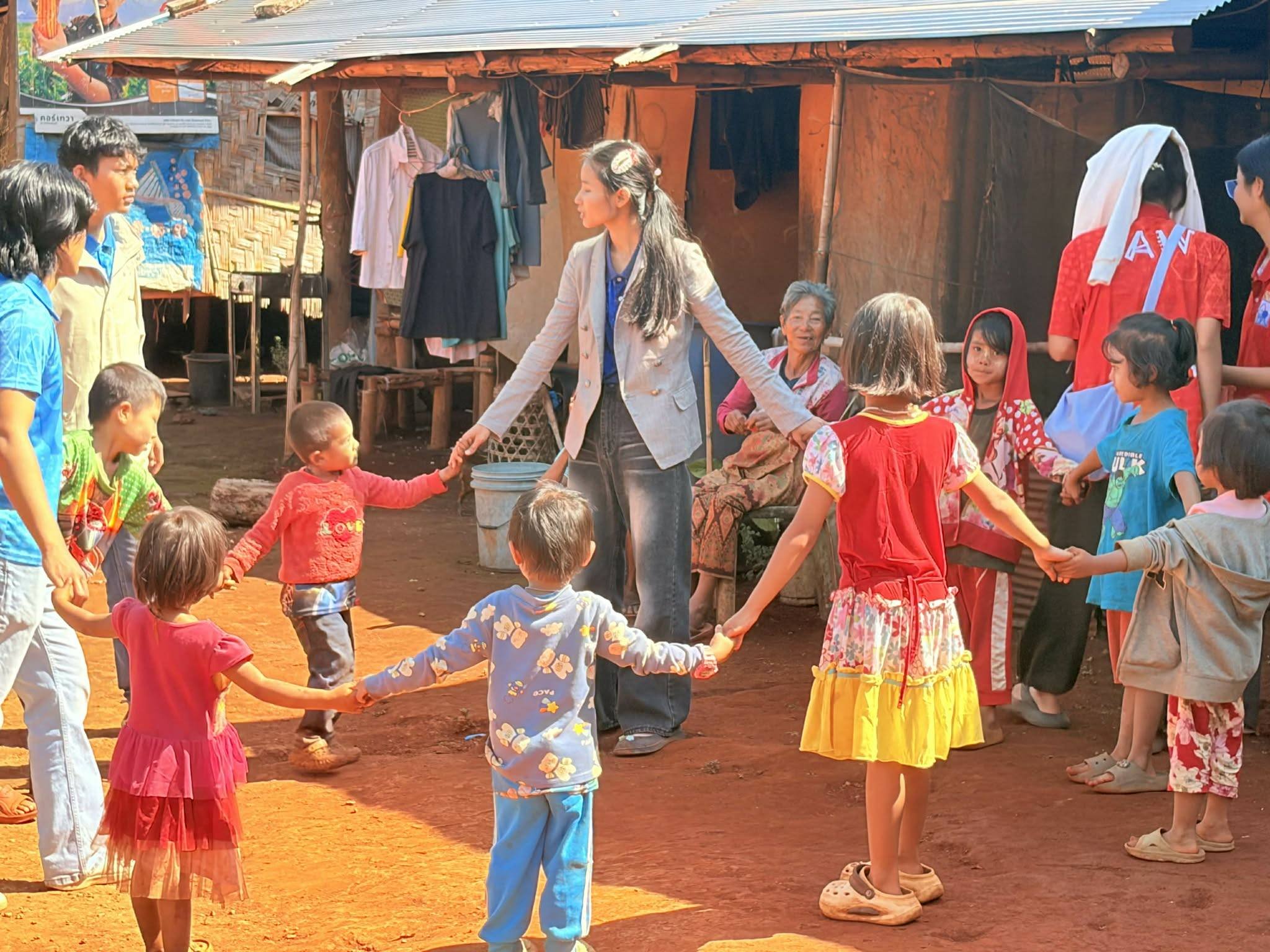 Group of children and women holding hands and playing in a circle outside in a rural area with a wooden house and hanging clothes in the background.