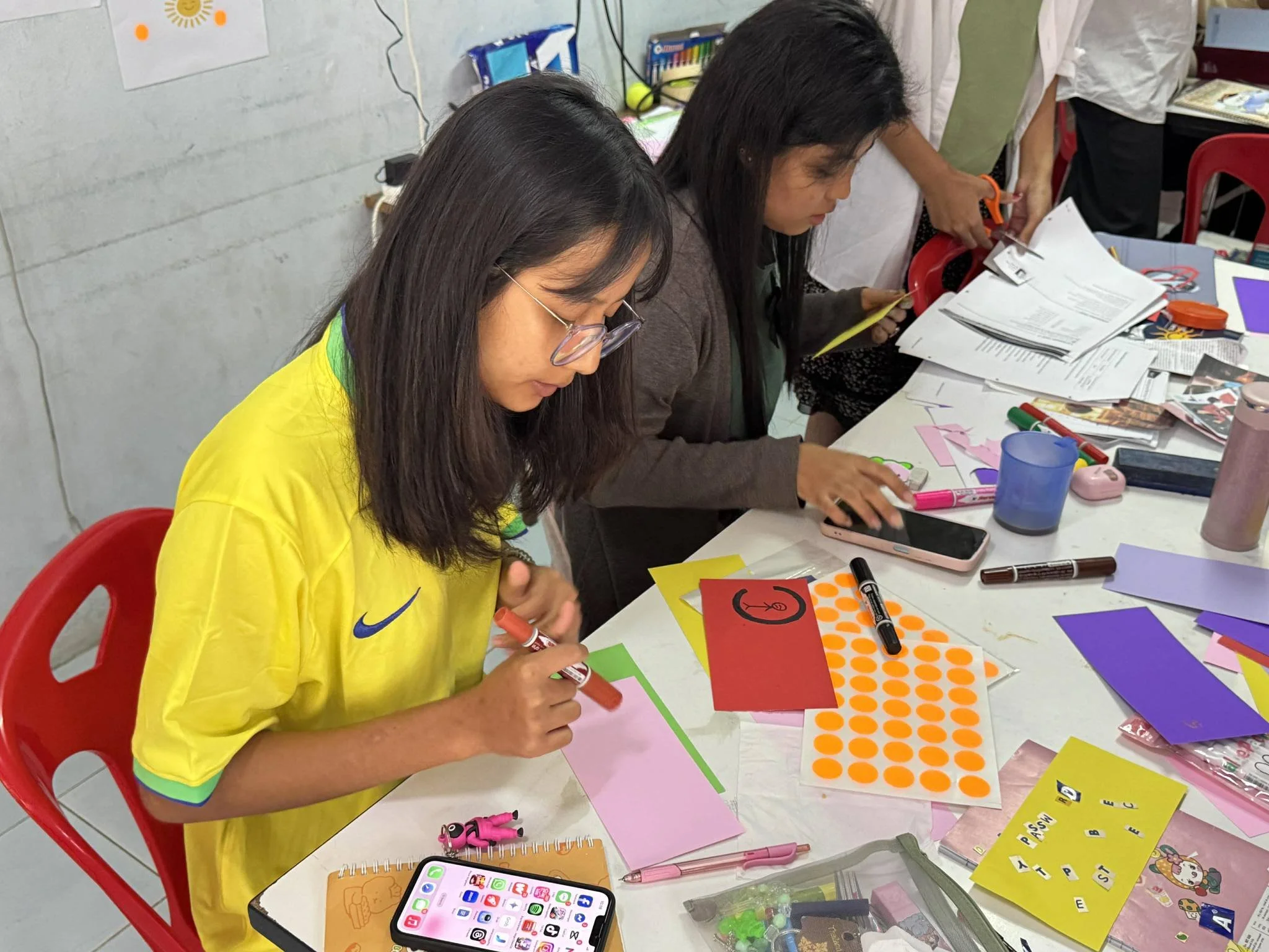 Two women engaged in arts and crafts at a cluttered table, working with colorful paper, markers, and stationery.
