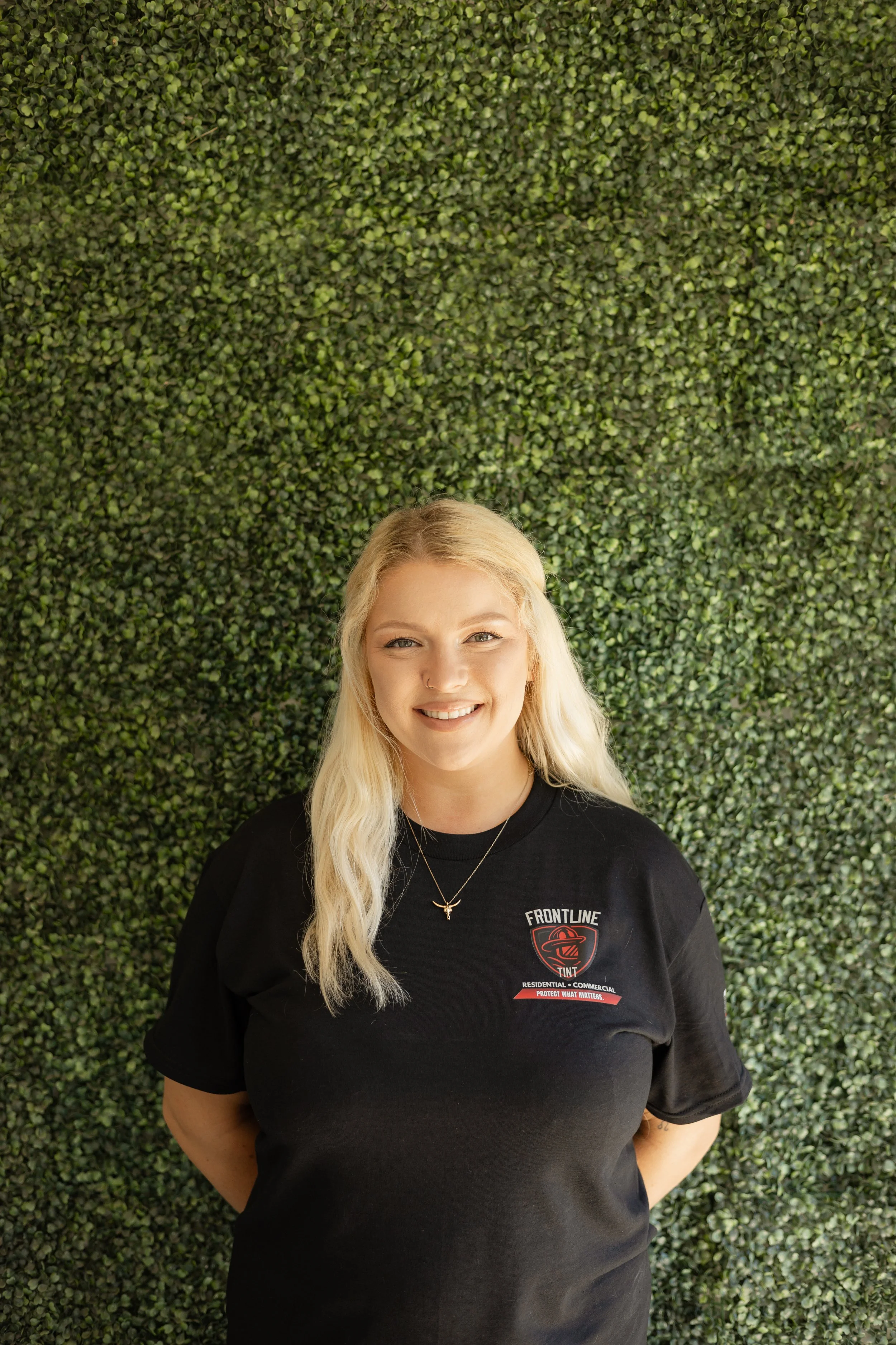 A smiling woman with long blonde hair wearing a black T-shirt with a logo that says "Frontline" and a shield emblem, standing in front of a large green leafy wall.