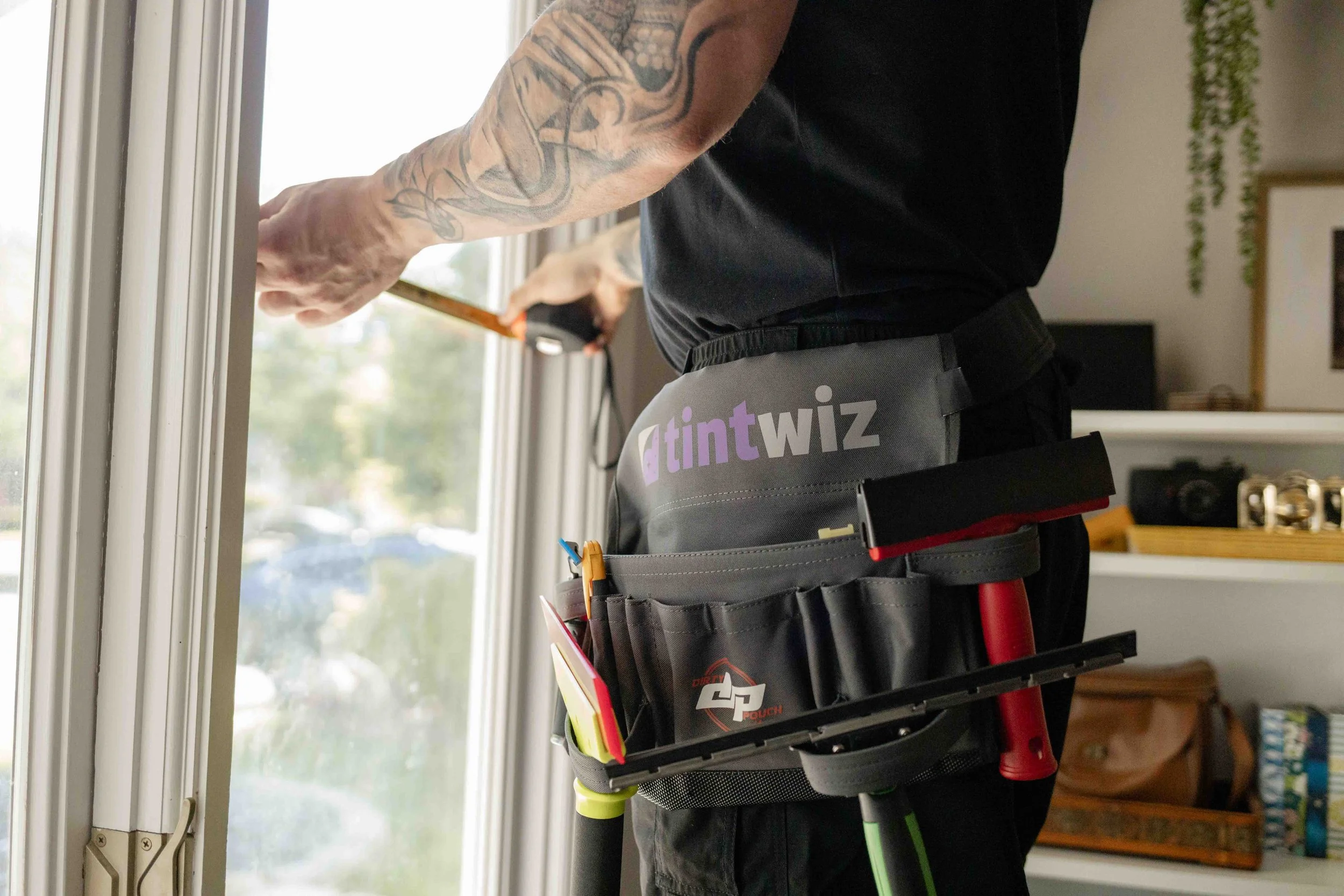 A person wearing black clothing and tool holster standing at a window, with tubbing and tools in the tool holder. In the background, a living room with a TV, bookshelf, and decorative plants.