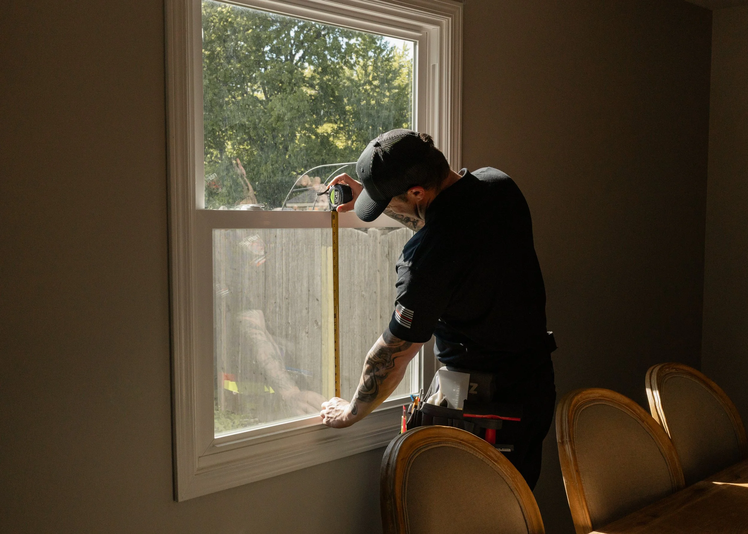 A man wearing a gray cap and black shirt is measuring a window with a yellow tape measure inside a room. There are chairs with wooden backs in the foreground, and outside the window, trees and a fence are visible.