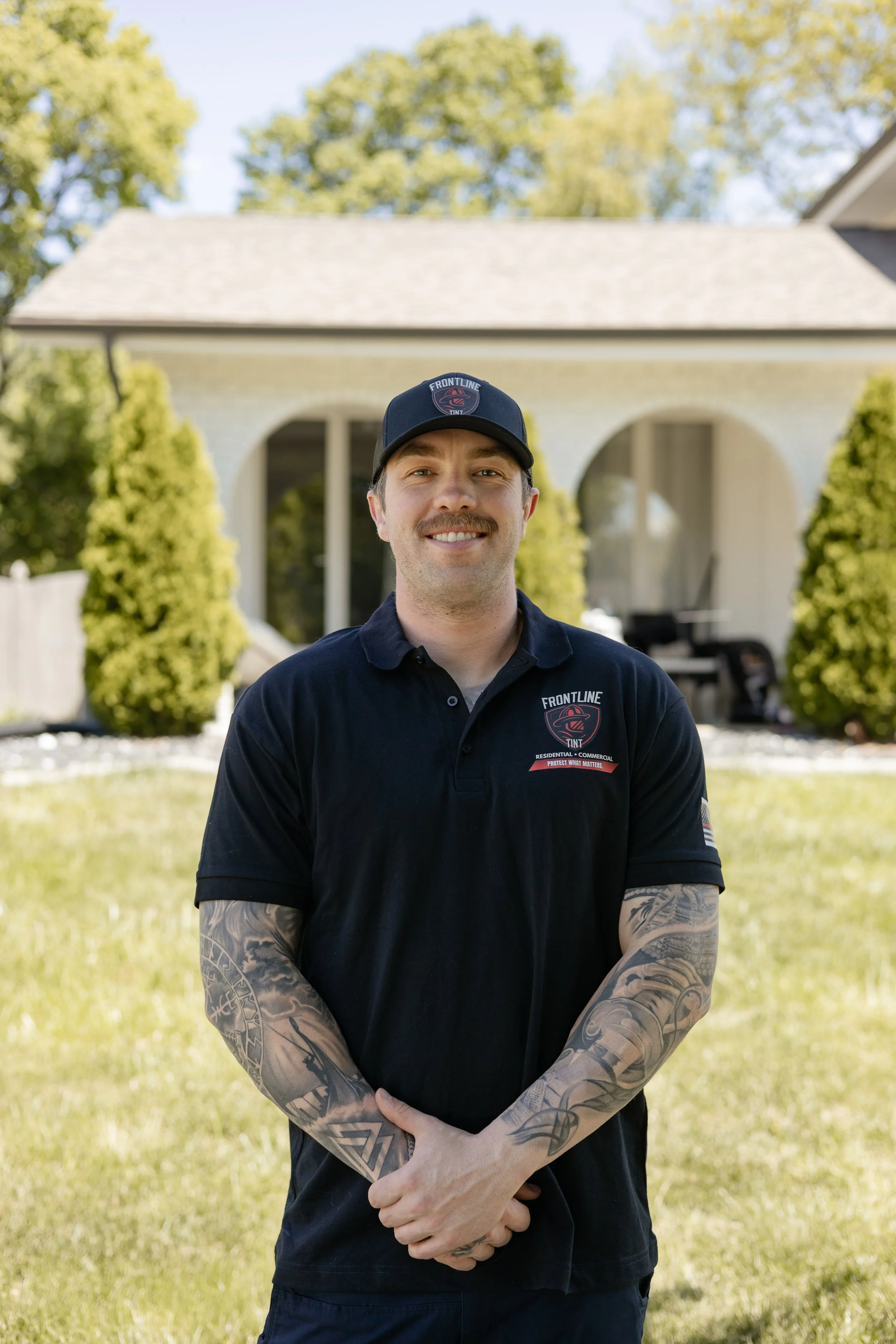 A man wearing a black polo shirt and a baseball cap with a logo, standing outdoors with house and trees in the background, smiling and showing tattoos on his arms.