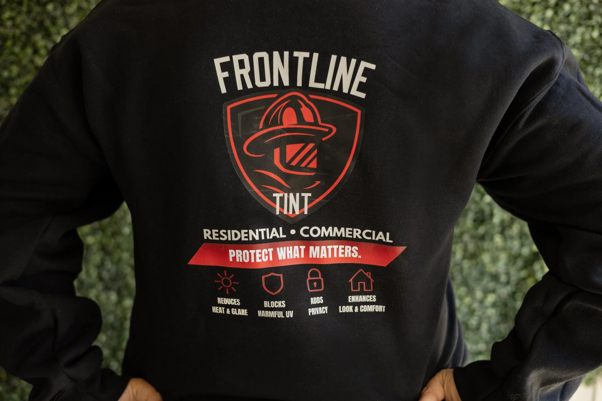 Person wearing a black jacket with a red and white logo for 'Frontline Tinte' and text indicating the company's services for residential and commercial properties, including protection against heat, glare, UV, and enhancing comfort.