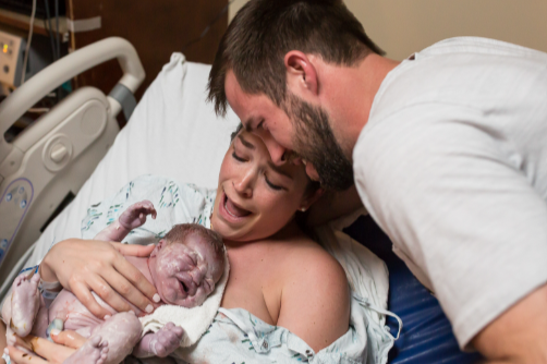 Newborn baby lying on hospital bed, woman in labor holding the baby, man leaning over them in a hospital room.