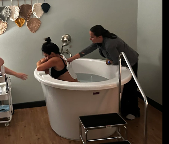 A woman helping a young girl into a round baptismal tub in a room with wooden flooring and decorative wall hangings.