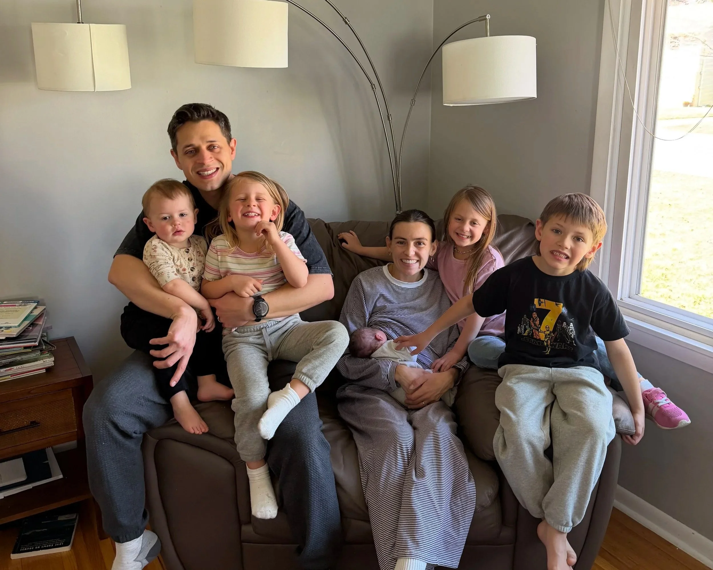Family sitting on a brown couch in a living room, smiling and posing for a photo. There are five children, an adult male, and a woman. One child is holding a newborn baby. Light streaming in from a large window on the right.