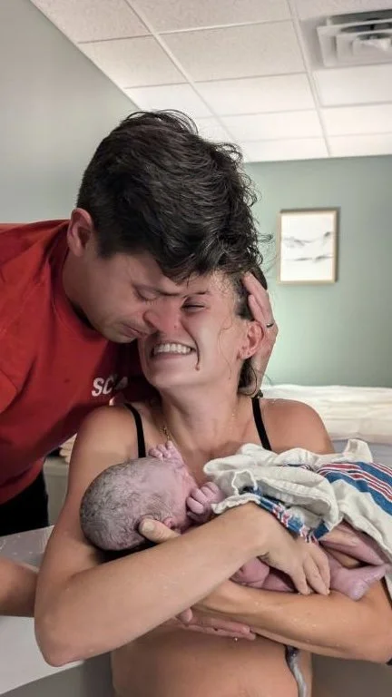 A woman holding a newborn baby in her arms, crying tears of joy, with a man leaning in to her in a hospital room.
