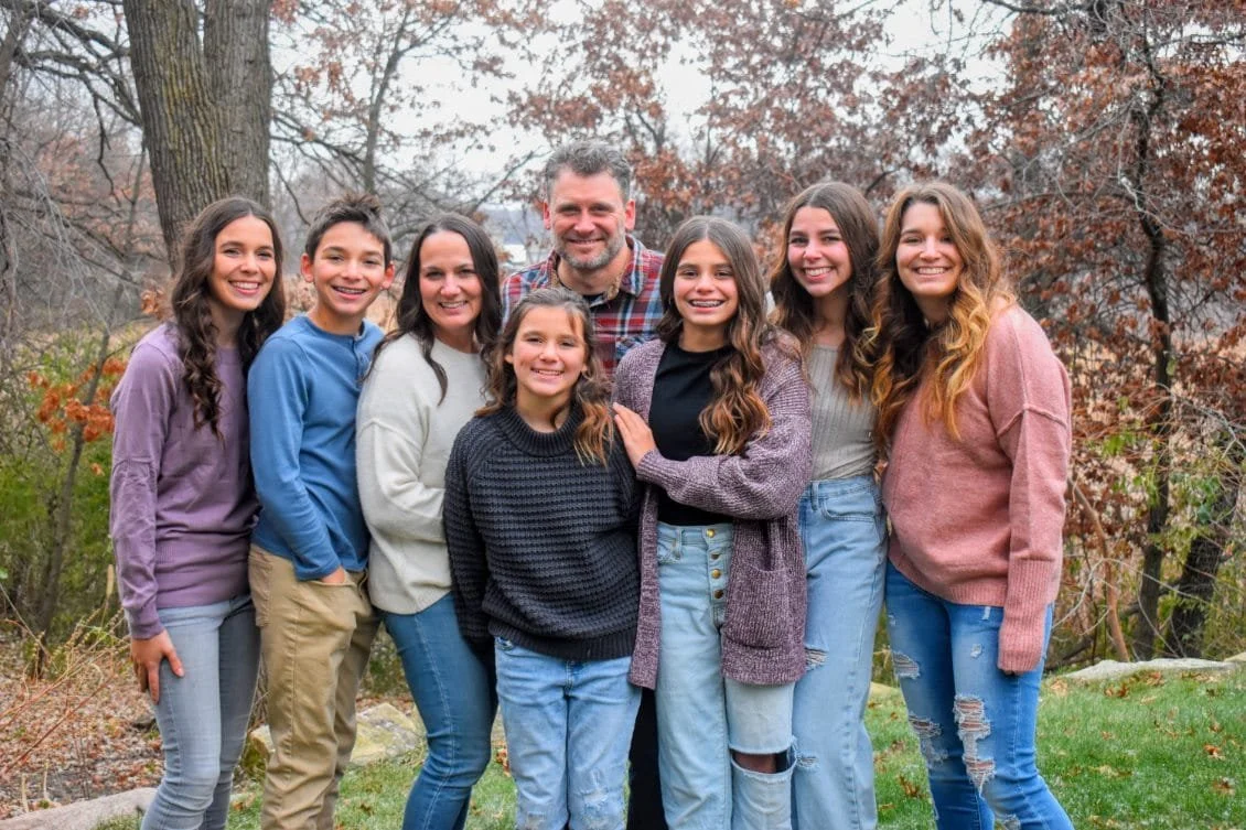 Family of nine posing outdoors in fall with trees in the background.