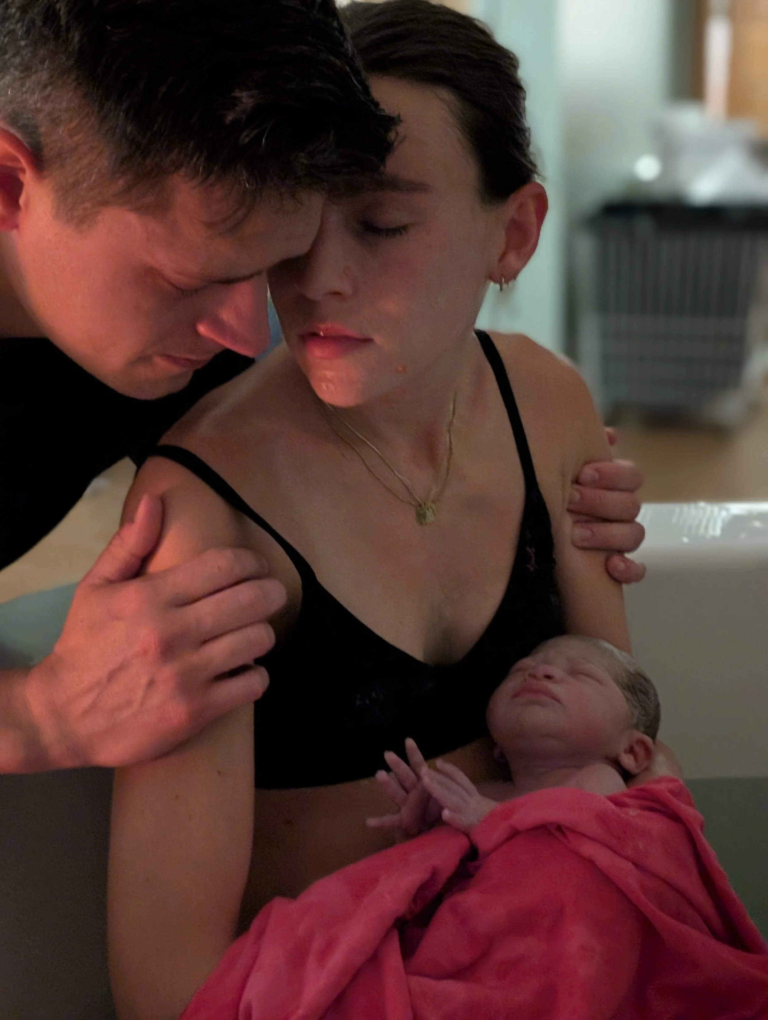 A woman holds a newborn baby wrapped in a pink blanket, while a man gently touches her shoulder, all in an indoor setting.